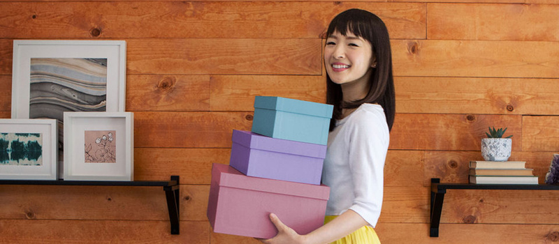 Marie Kondo sonríe y carga tres cajas de colores frente a una pared de madera con estantes y cuadros